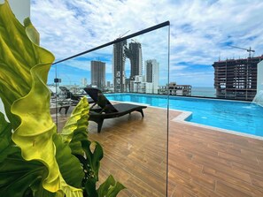 A rooftop pool with a city skyline view, lounge chairs, and a wooden deck.