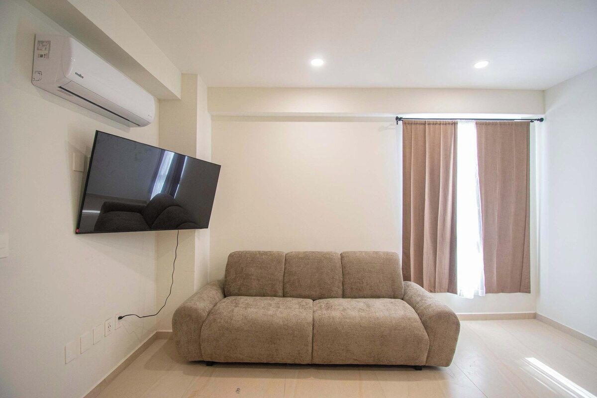A living room with a flat-screen TV mounted on the wall, a beige sofa, and a window with curtains.