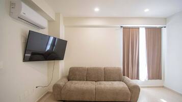 A living room with a flat-screen TV mounted on the wall, a beige sofa, and a window with curtains.
