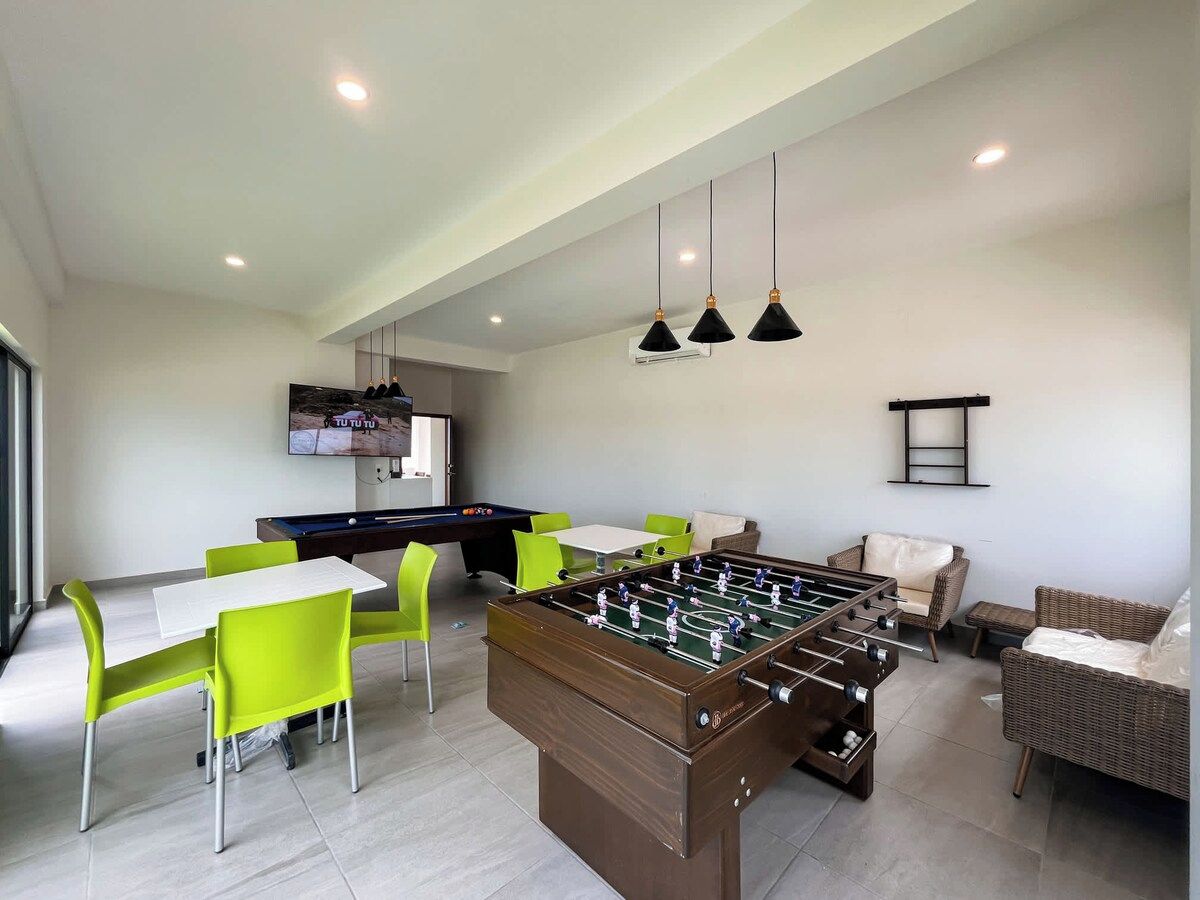A modern living room with a foosball table, bright green chairs, a pool table, and a flat-screen TV.