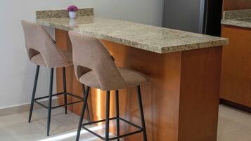 A kitchen with a granite countertop, two bar stools, a stainless steel refrigerator, and a potted plant on the counter.