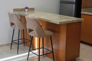 A kitchen with a granite countertop, two bar stools, a stainless steel refrigerator, and a potted plant on the counter.