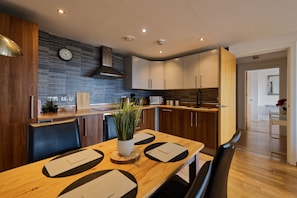 A modern kitchen with wooden cabinets, a black backsplash, and a dining area with a wooden table and chairs. - L1 Penthouse with Private Balcony and Views (Liverpool)