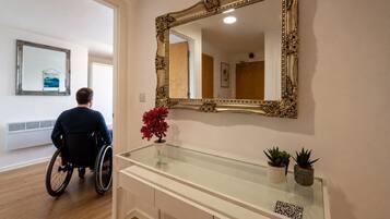 A wheelchair-accessible room with a large ornate mirror, a glass-top console table with potted plants, and a view of a hallway.