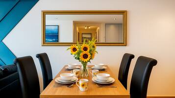 A dining area with a wooden table, black chairs, white plates, bowls, and a vase of sunflowers. A large mirror hangs on the wall above the table.