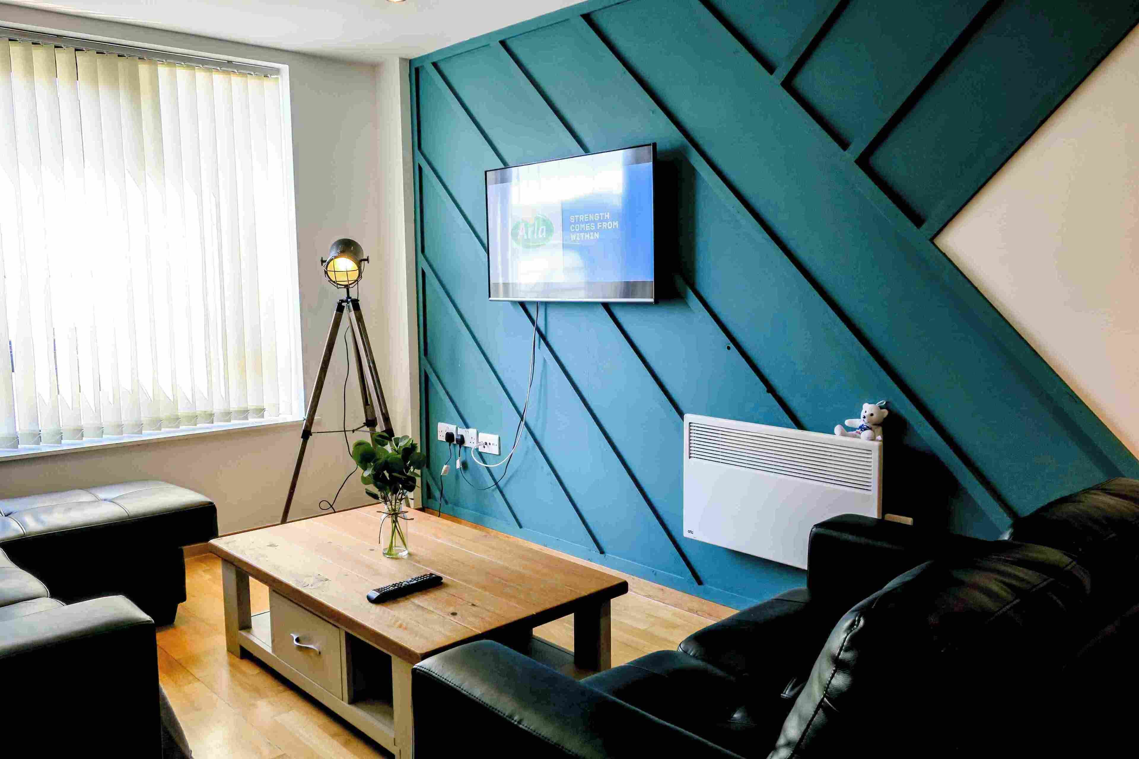 A modern living room with a teal accent wall, a mounted television, a tripod lamp, a wooden coffee table, and a black leather sofa.