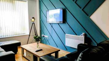A modern living room with a teal accent wall, a mounted television, a tripod lamp, a wooden coffee table, and a black leather sofa.