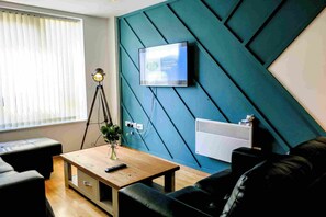 A modern living room with a teal accent wall, a mounted television, a tripod lamp, a wooden coffee table, and a black leather sofa. - Central Access, Self-Check-in, 2BR2Bath, Stepless (Liverpool)