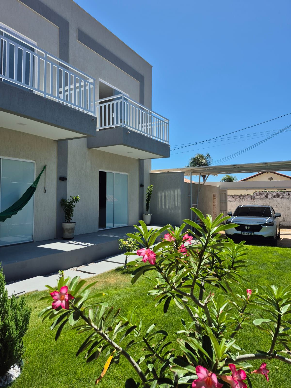 A modern two-story house with a balcony, a car parked in the driveway, and a well-maintained lawn with flowering plants.
