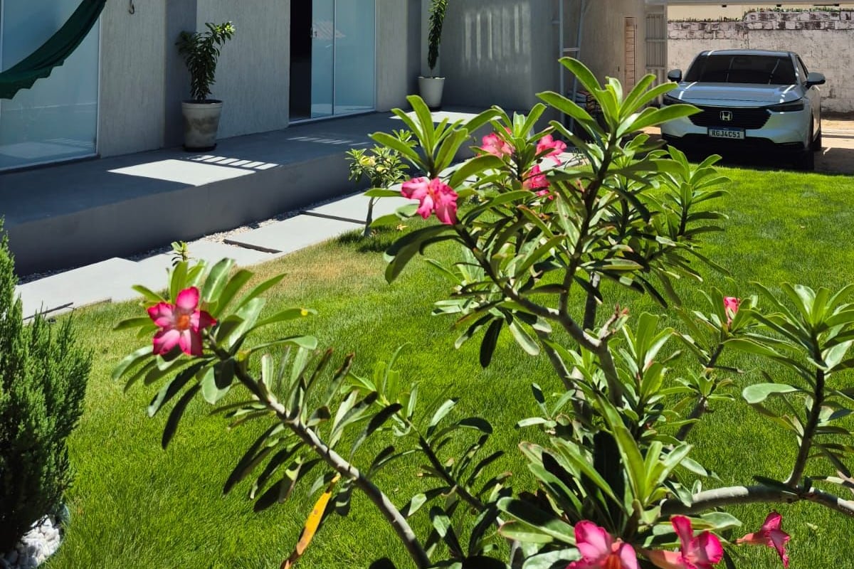A modern two-story house with a balcony, a car parked in the driveway, and a well-maintained lawn with flowering plants.
