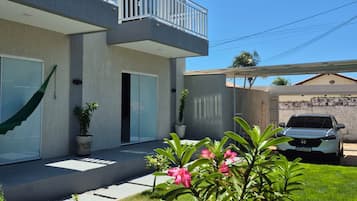 A modern two-story house with a balcony, a car parked in the driveway, and a well-maintained lawn with flowering plants.