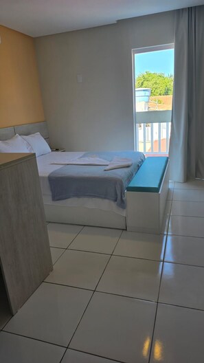 A hotel room with a bed, a bench, a desk, and a window with a view of greenery outside.