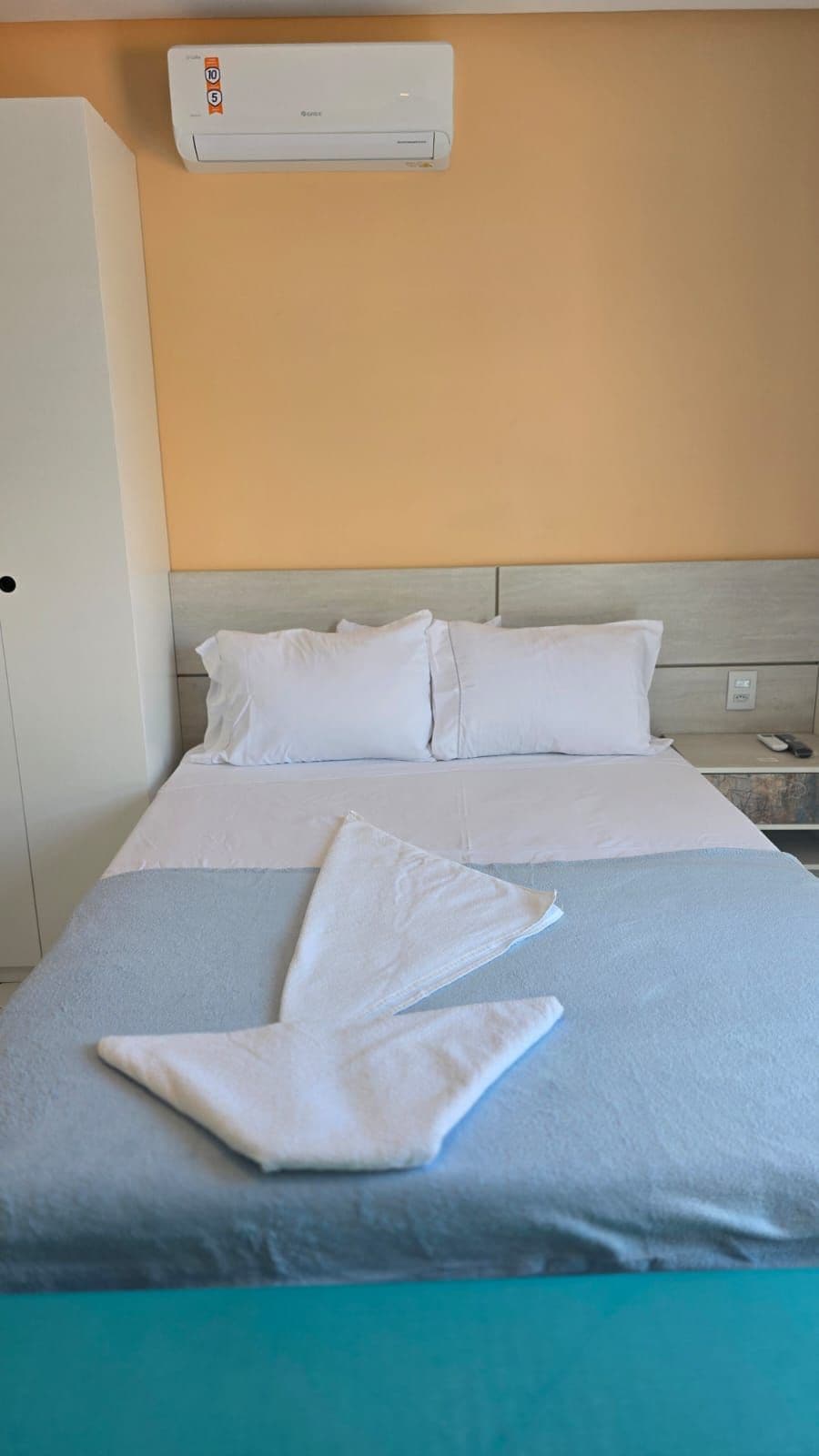 A hotel room with a bed, pillows, a blanket, an air conditioning unit, and a nightstand.