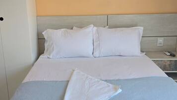 A hotel room with a bed, pillows, a blanket, an air conditioning unit, and a nightstand.