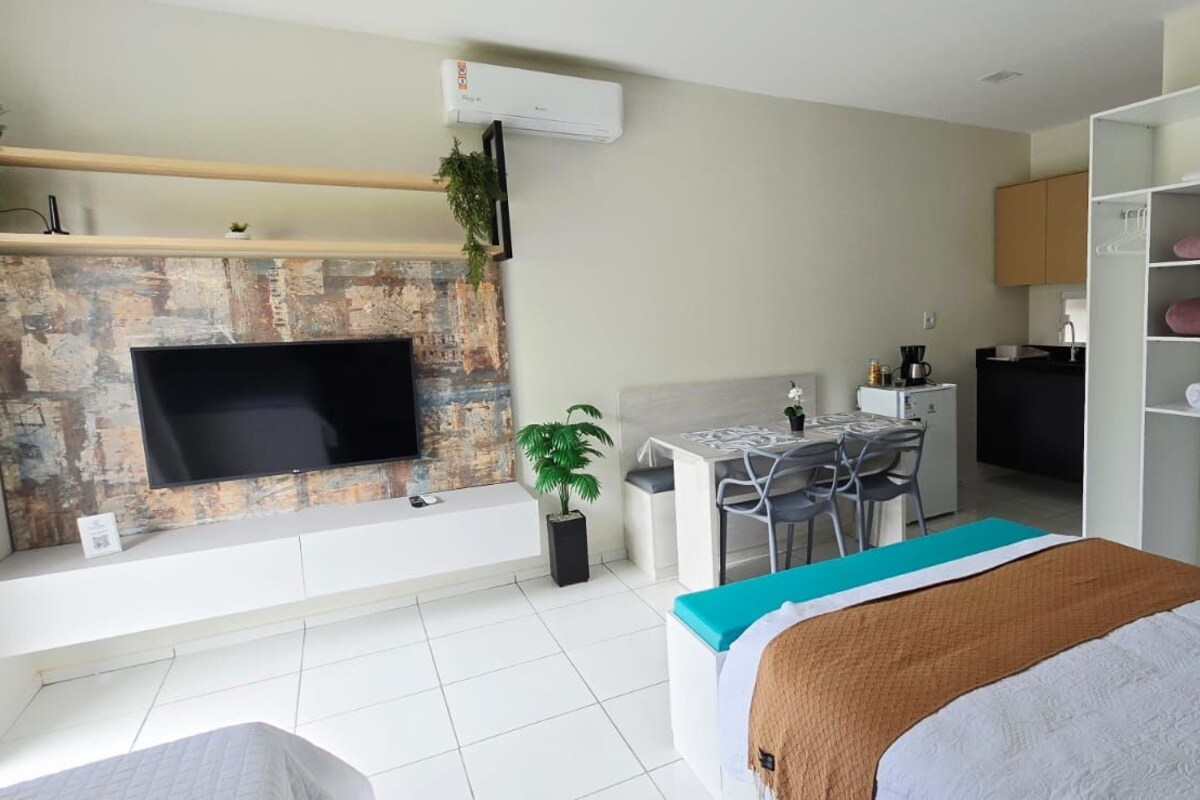 Habitación de hotel moderna con televisor de pantalla plana montado en una pared con textura, un escritorio con sillas, una cama con manta azul y marrón, y un armario con estantes.