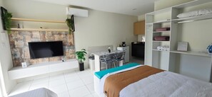 Een moderne hotelkamer met een flatscreen televisie aan een gestructureerde wand, een bureau met stoelen, een bed met een blauw-bruine deken en een kledingkast met planken.