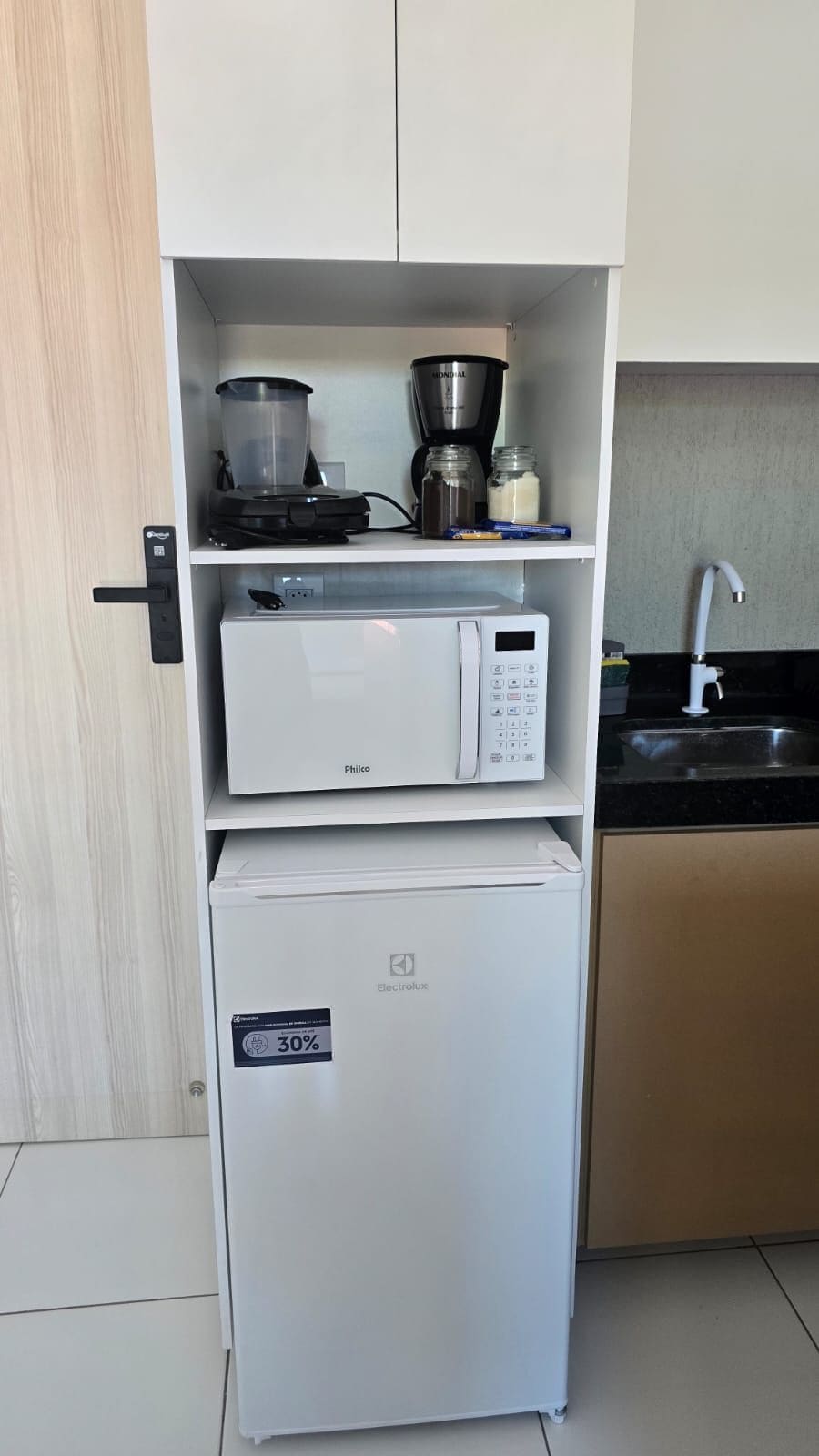 Cozinha compacta com micro-ondas embutido, pequena geladeira e máquina de café no balcão.