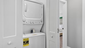 A compact, white laundry room with a built-in washer and dryer, a small shelf with a bottle, and a caution sign. - Riverside House by Avantstay French Broad River Views Balcony Near DT + Silver Line Park (Asheville)