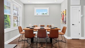 Comedor con una mesa rectangular dispuesta para seis personas, rodeada de seis sillas, una ventana con persianas, una puerta y un cuadro en la pared.