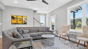 A modern living room with a grey sofa, two armchairs, a coffee table, and a staircase. - Riverside House by Avantstay French Broad River Views Balcony Near DT + Silver Line Park (Asheville)