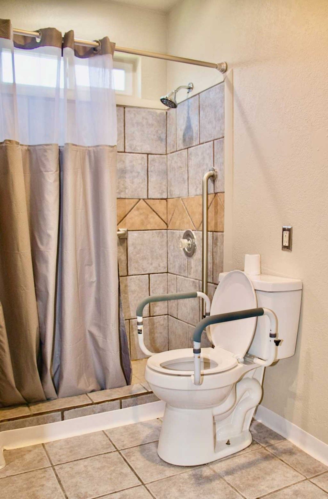 A bathroom with a shower, a toilet with a grab bar, and a window with a curtain.