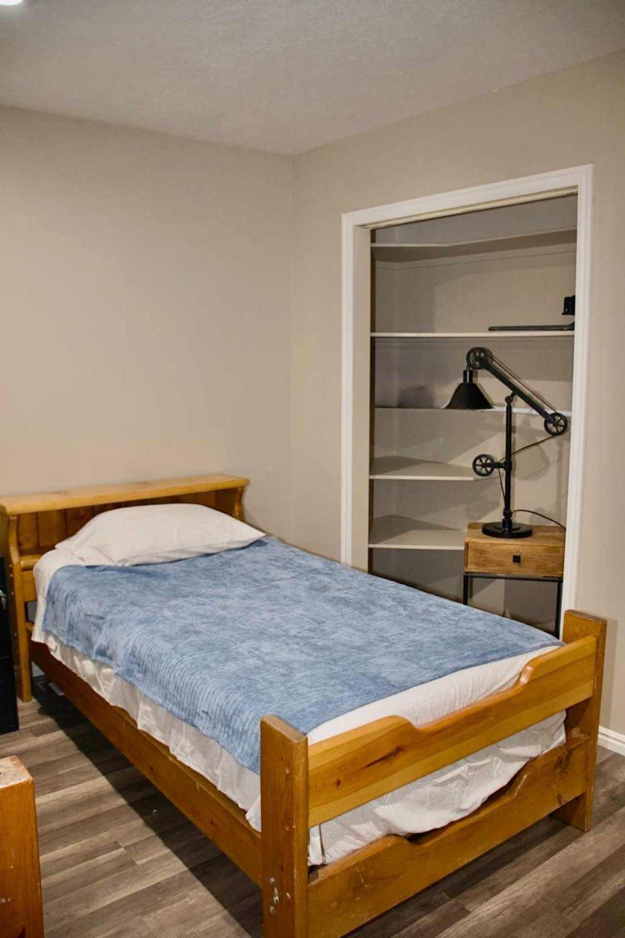 A single bed with a wooden frame and a blue blanket, a bedside table with a lamp, and a wardrobe with shelves.