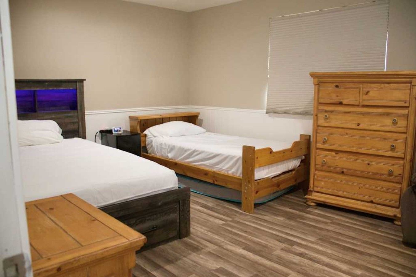 A bedroom with two single beds, a wooden dresser, a nightstand, and a window with blinds.
