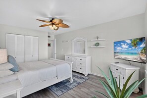 Une chambre à coucher avec un lit, un ventilateur de plafond, une télévision et une fenêtre donnant sur une plage.