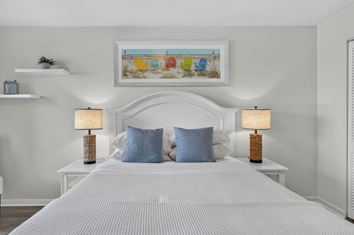 Une chambre avec un lit blanc, deux tables de chevet avec des lampes, une étagère murale ornée d’une plante et un tableau encadré au-dessus du lit.