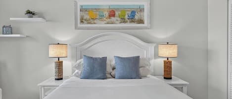 Une chambre avec un lit blanc, deux tables de chevet avec des lampes, une étagère murale ornée d’une plante et un tableau encadré au-dessus du lit.