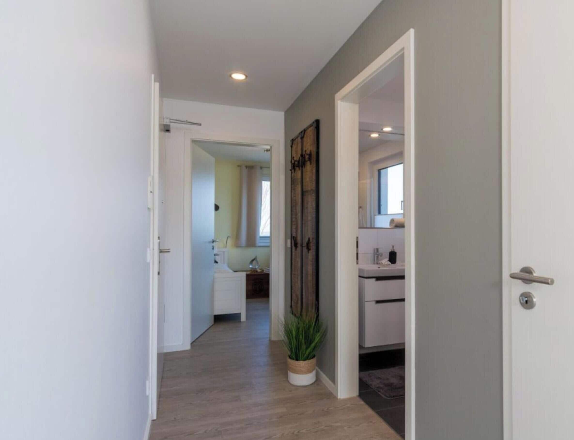 A hallway with a plant, a door leading to a bathroom, and a view into a room with a window.