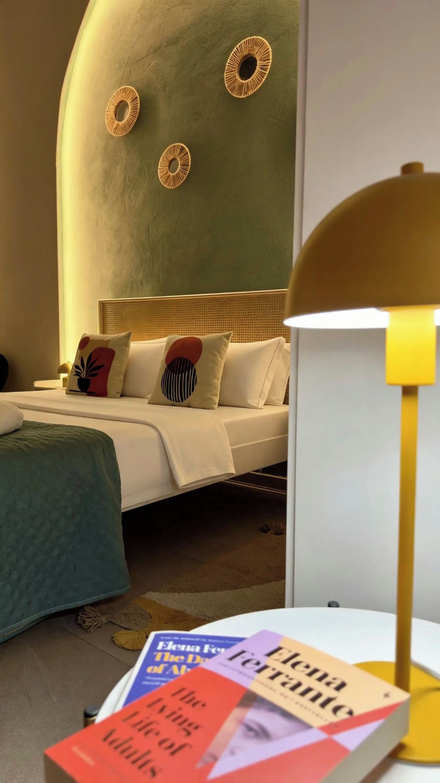 A hotel room with a bed, pillows, a bedside table with books, and a lamp.