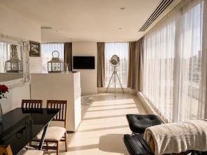 Ein modernes Wohnzimmer mit Essbereich, einem Flachbildfernseher, einem Stativ und großen Fenstern mit Vorhängen.