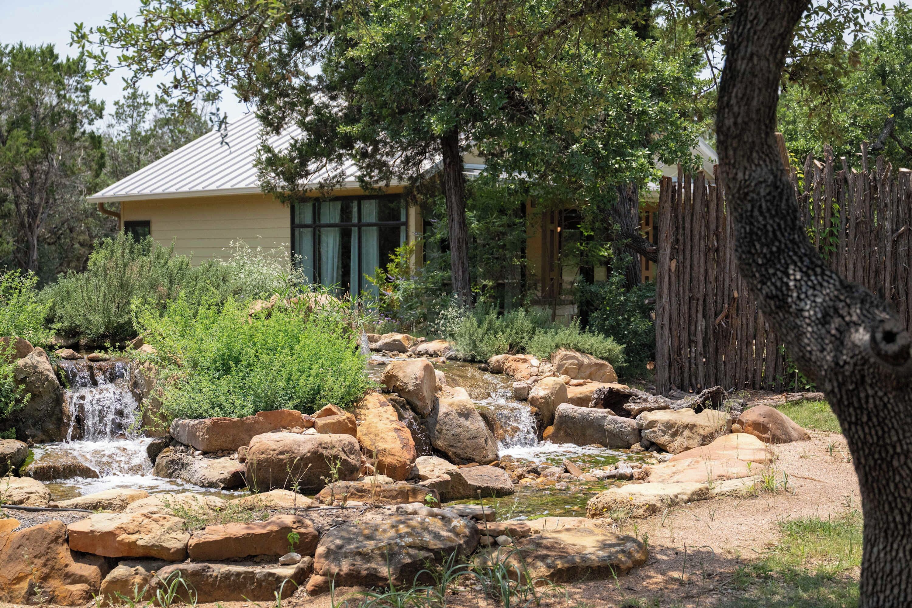A rustic outdoor setting with a house, a wooden fence, and a natural landscape with rocks and a small waterfall.