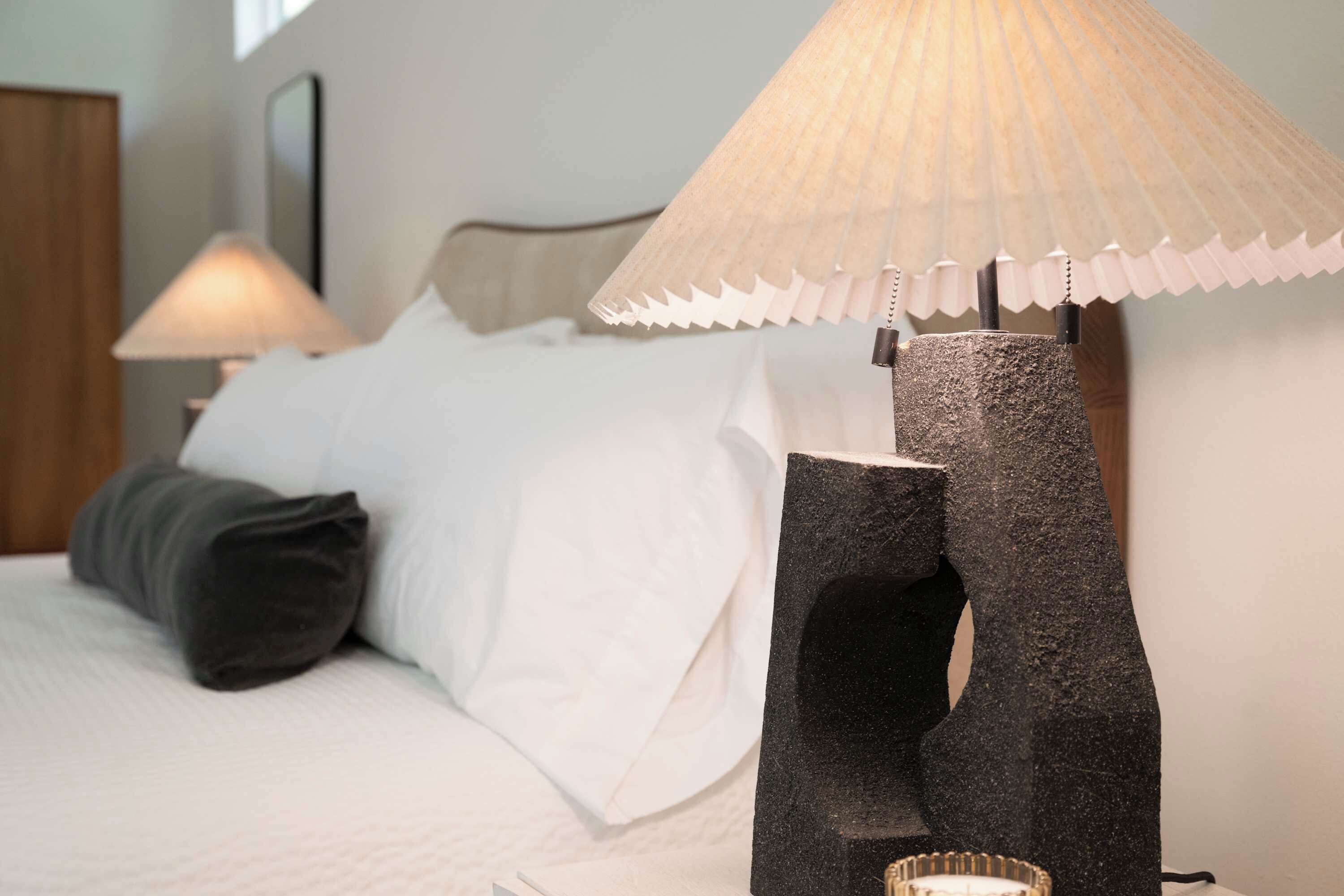 A neatly made bed with white linens and a dark pillow, a modern lamp with a textured shade, and a wall-mounted mirror.