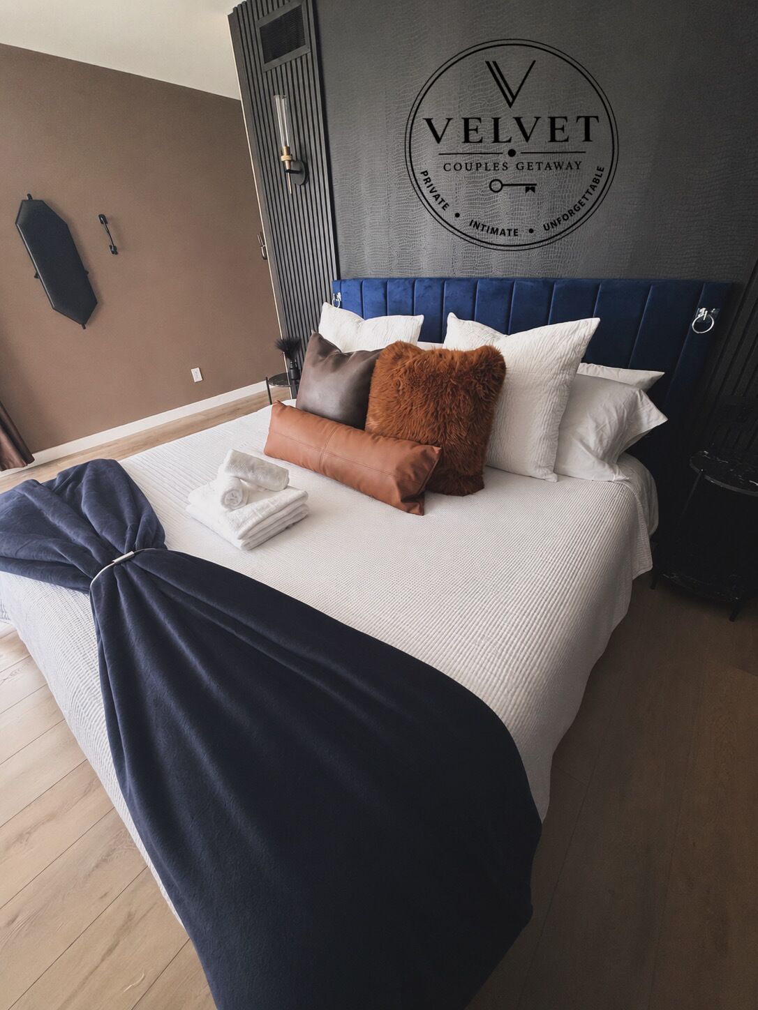 Una cama bien hecha con cabecera azul, un logotipo en la pared y una mesita de noche con lámpara.