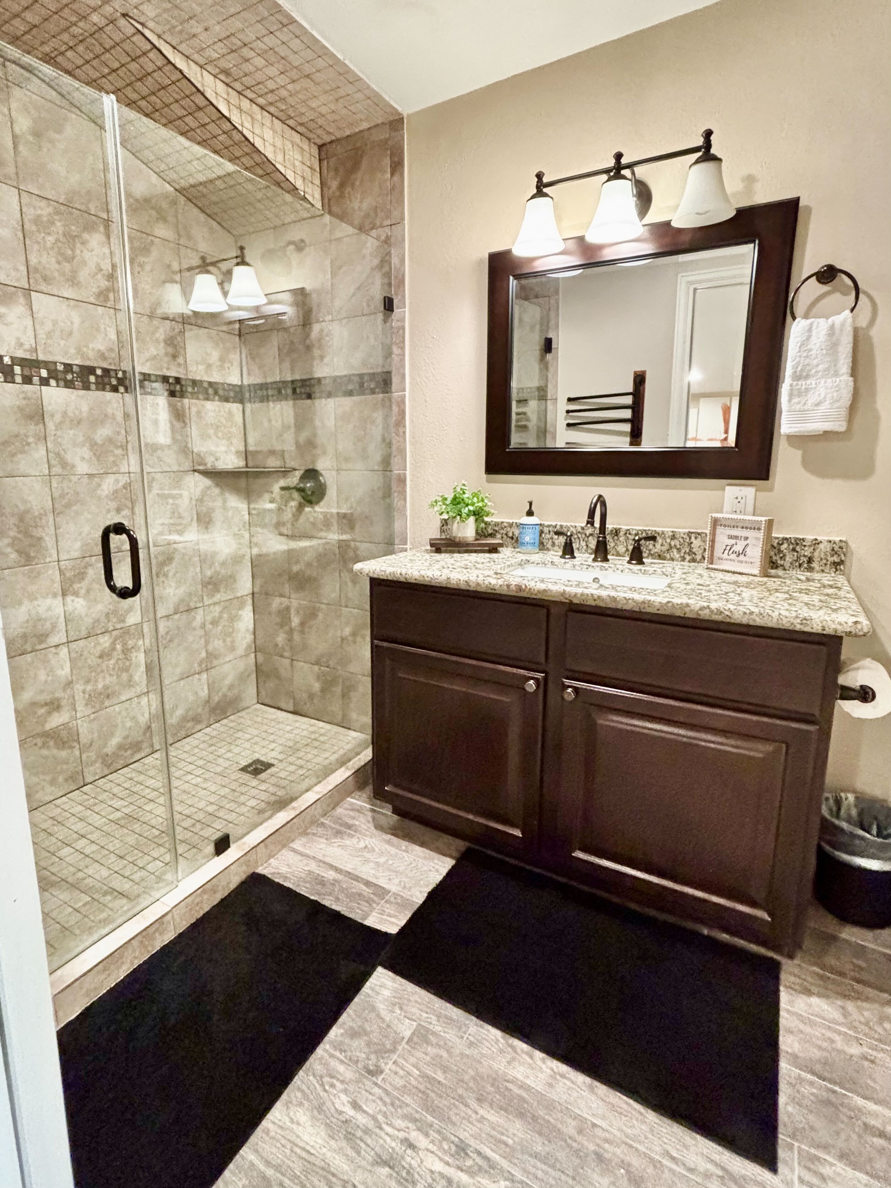 A bathroom with a glass-enclosed shower, a vanity with dark cabinets, a mirror with three lights, and a towel rack.