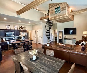 A spacious living area with a wooden loft, a dining table with chairs, a leather sofa, and a TV mounted on the wall. - The Vine (New Ulm)
