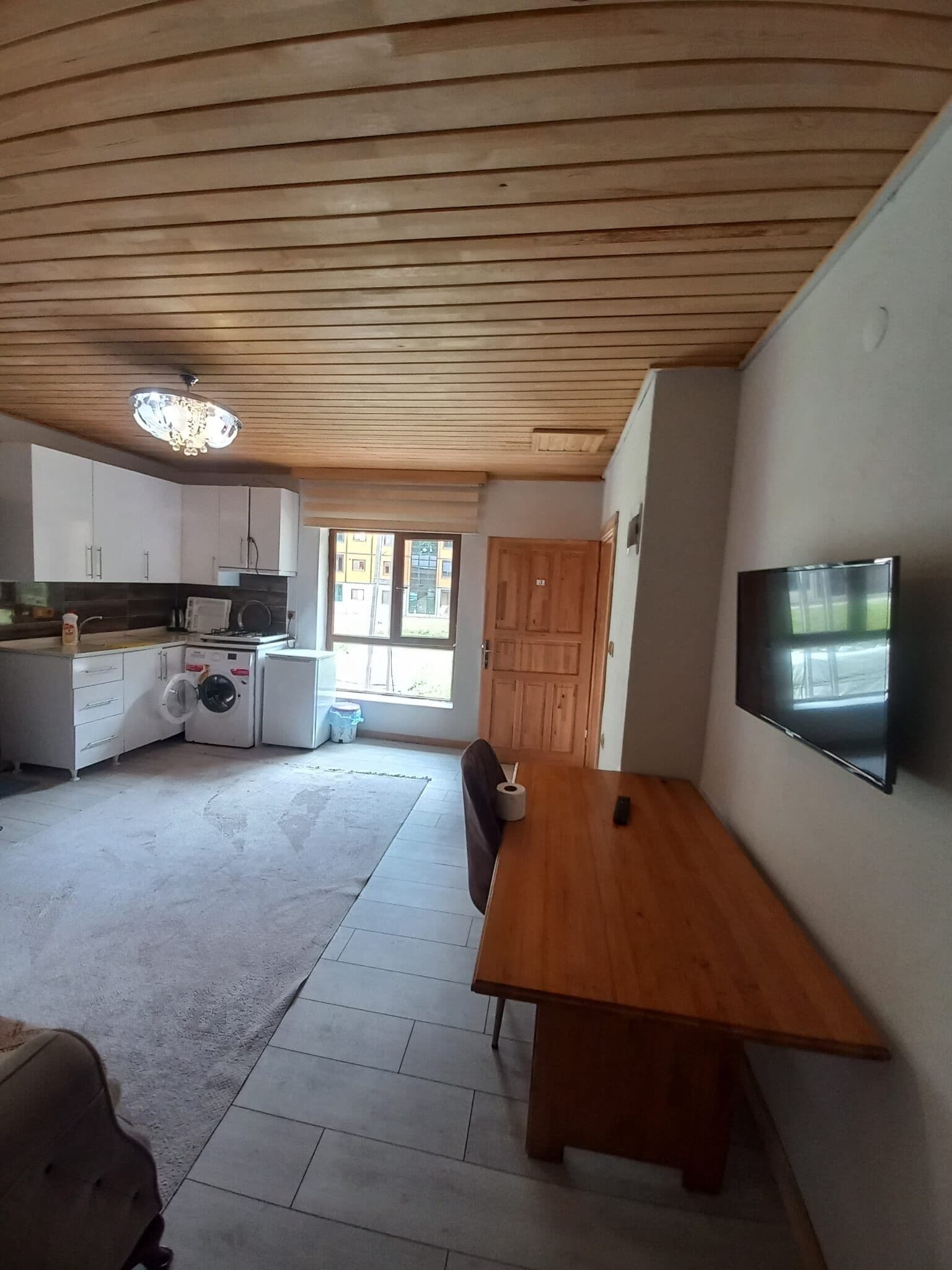 Ein geräumiger Wohnbereich mit einem Holzessttisch, einem an der Wand befestigten Fernseher und einer Küche mit modernen Geräten.