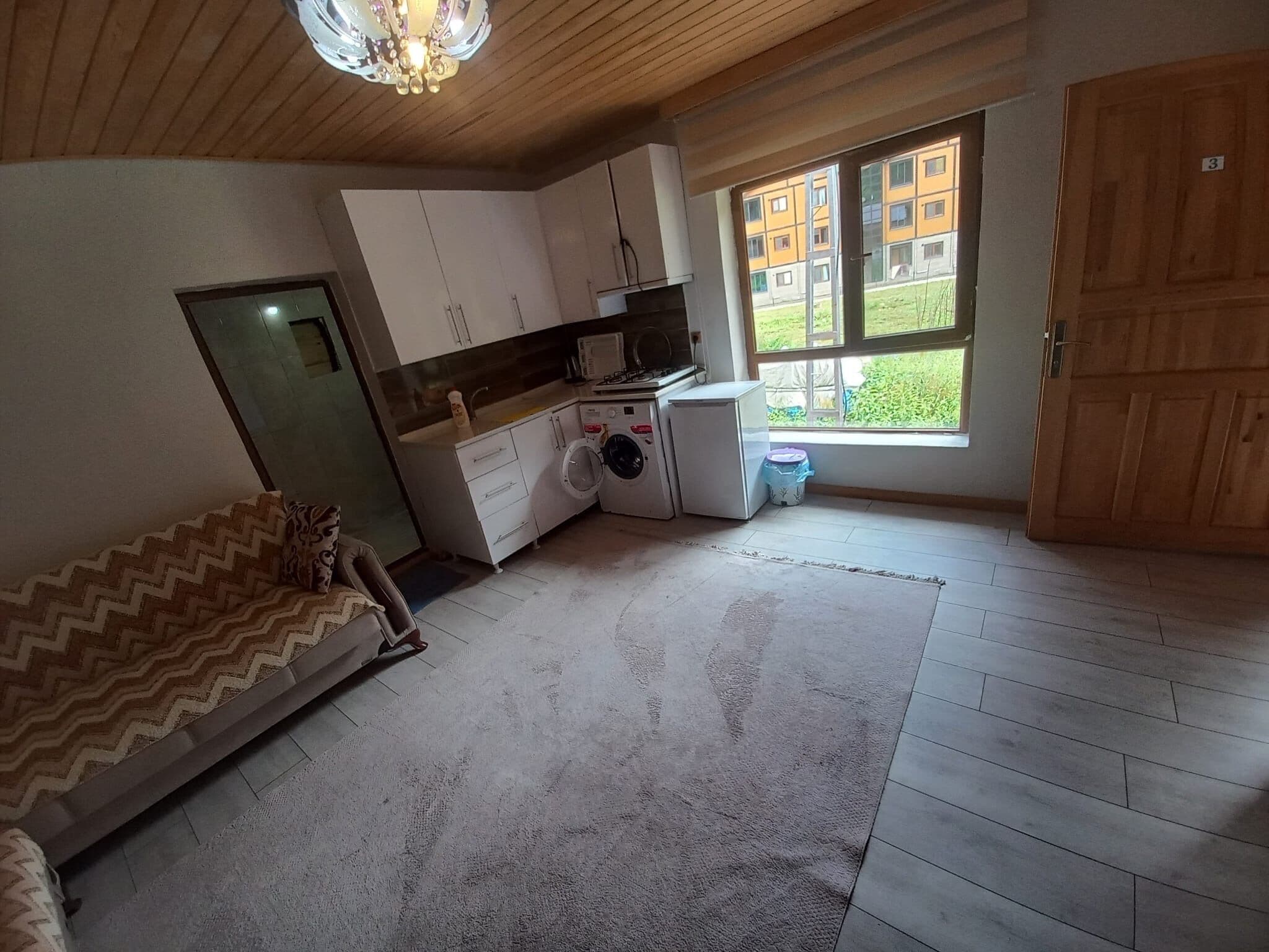 Habitación pequeña con cama, lavadora, microondas y ventana con vistas a edificios.
