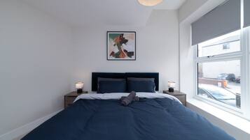 Une chambre avec un grand lit, deux tables de chevet, un luminaire au plafond, une fenêtre donnant sur l’extérieur et un tableau abstrait encadré accroché au mur.