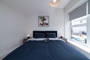 Ein Schlafzimmer mit einem großen Bett, zwei Nachttischen, einer Deckenleuchte, einem Fenster mit Blick ins Freie und einem gerahmten abstrakten Gemälde an der Wand.
