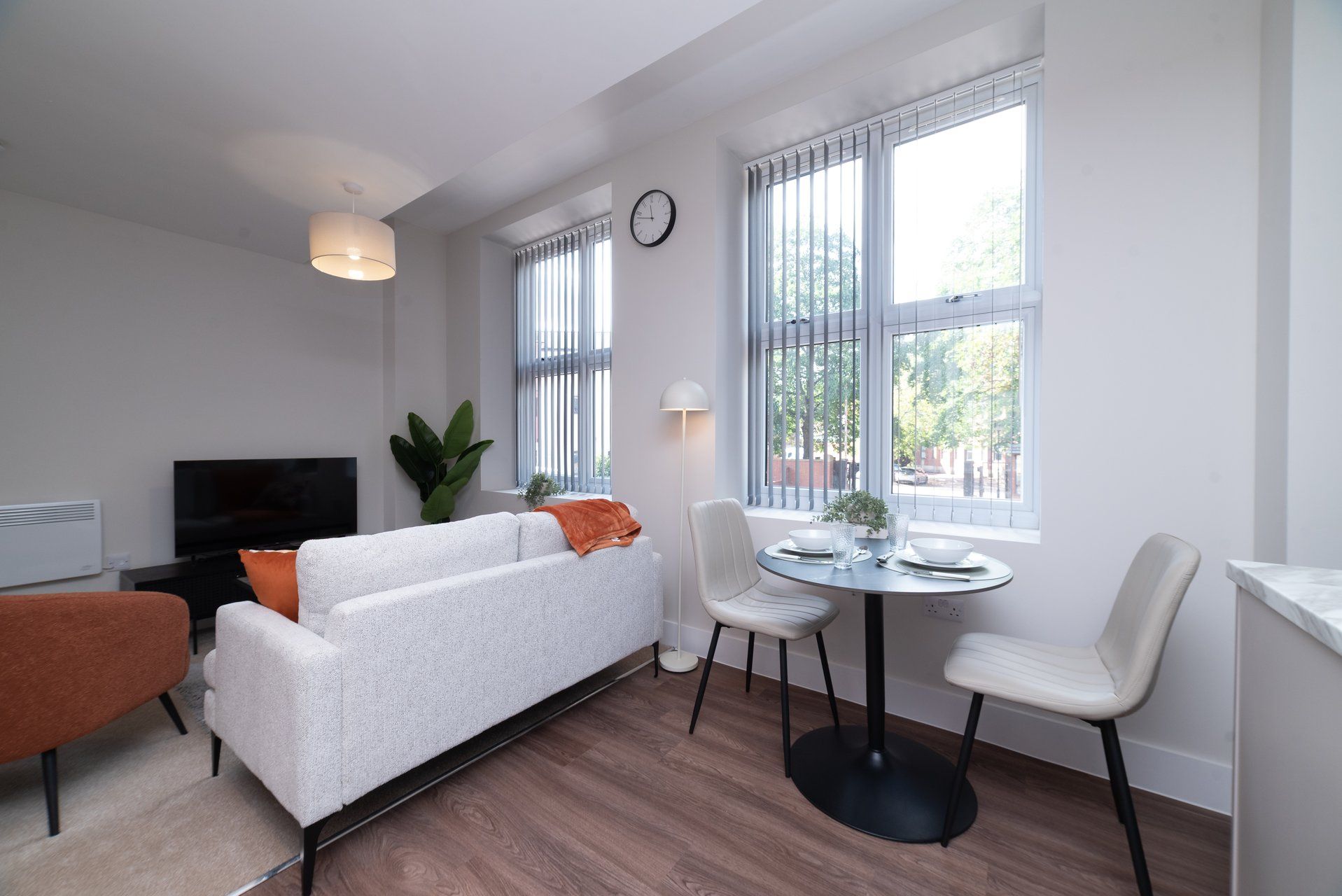 A modern living room with a sofa, a dining table with chairs, a TV, and a large window with blinds.