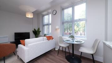 A modern living room with a sofa, a dining table with chairs, a TV, and a large window with blinds.