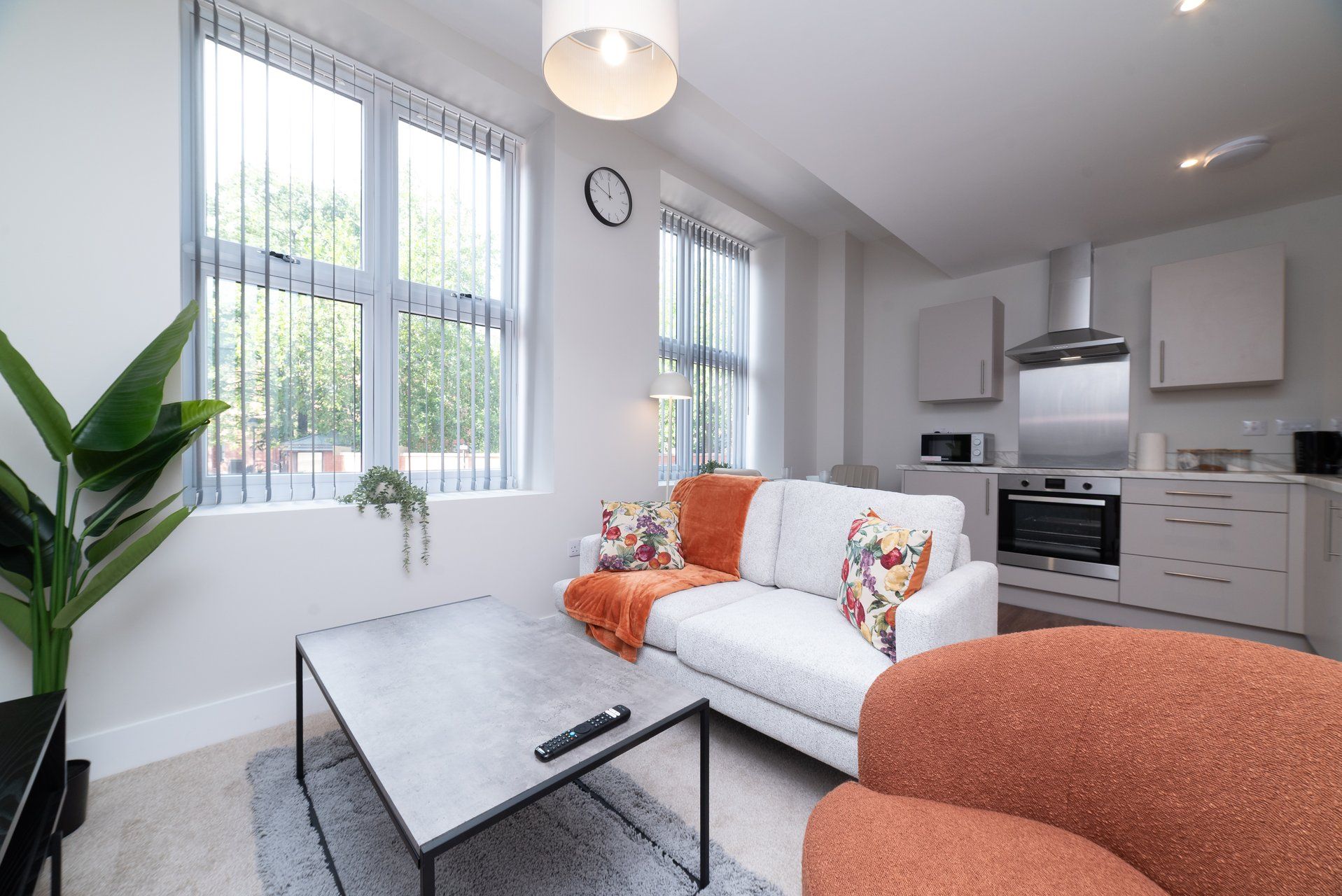 A modern living room with a sofa, a coffee table, and a TV. There is a kitchen area with a microwave, oven, and cabinets.