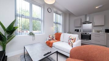 A modern living room with a sofa, a coffee table, and a TV. There is a kitchen area with a microwave, oven, and cabinets.