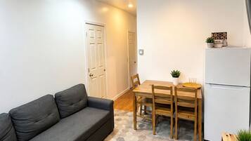A compact living space with a grey sofa, a wooden dining table with chairs, a white refrigerator, and a door leading to another room.