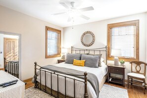 Un dormitorio con cama, mesitas de noche, una silla, ventilador de techo y ventana con persianas.