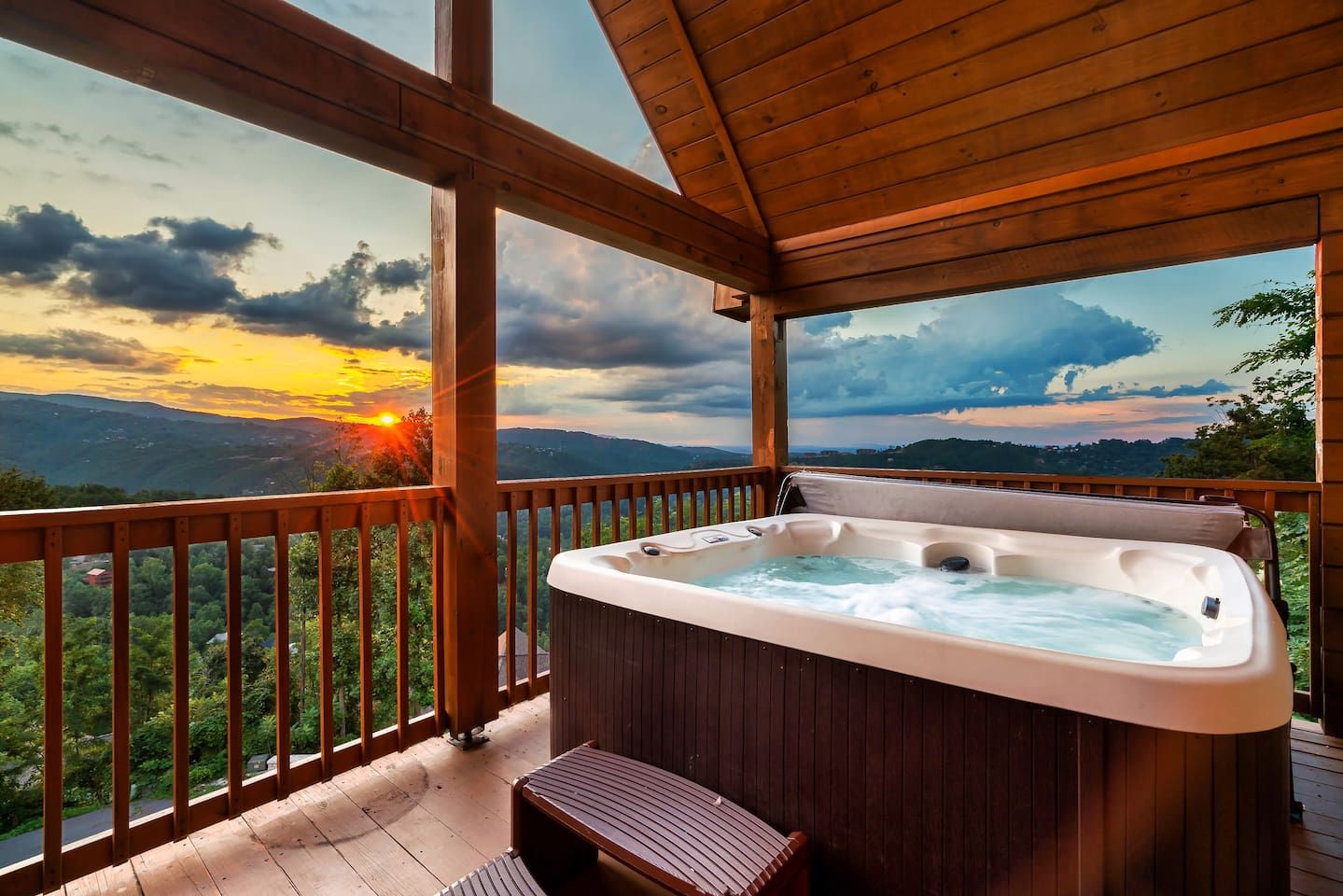 A wooden deck with a hot tub and a bench, overlooking a scenic landscape with mountains and a sunset.