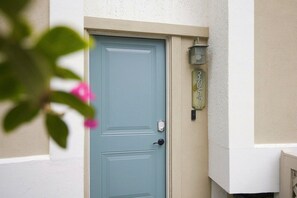 A blue door with a number plate, a wall-mounted light fixture, and a partial view of a plant with pink flowers. - MyStay at 3057 Seascape | South Forest Beach | Private Beach Cabana | Sleeps 7 (Hilton Head Island)